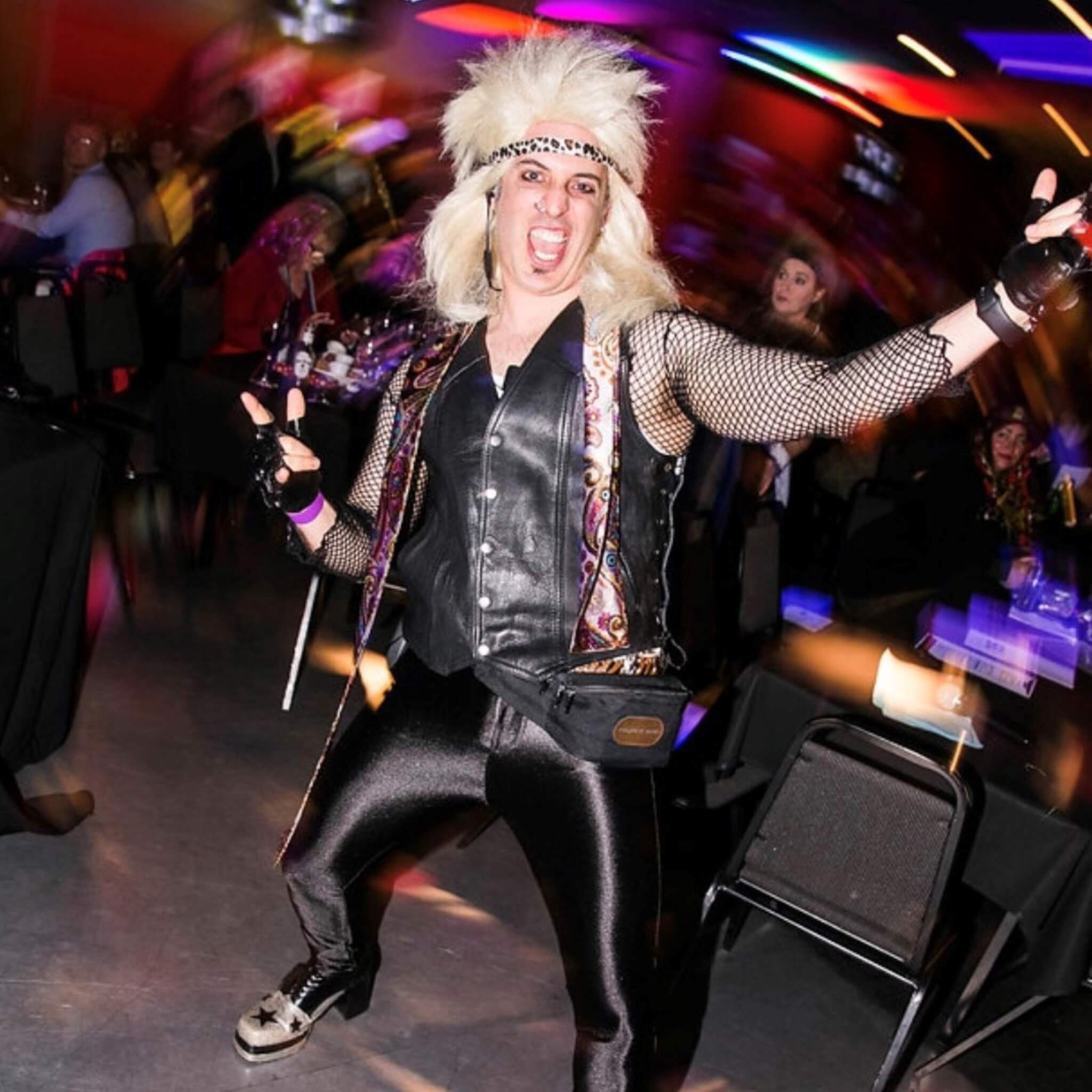 OneStopBox founder dressed in an 80s rock star costume with blonde wig, leather vest and fishnet sleeves, performing at a themed party event. Paulie is the ultimate Chief Party Officer