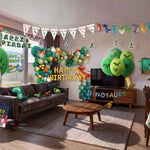 Complete dinosaur themed kids birthday party decorations filling a living room, including green and gold balloon garland, printed balloons, dinosaur foil balloons, hanging swirls, bunting, happy birthday sign, wall decal, light box and table cover from a OneStopBox all-in-one party kit.