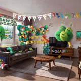 Complete dinosaur themed kids birthday party decorations filling a living room, including green and gold balloon garland, printed balloons, dinosaur foil balloons, hanging swirls, bunting, happy birthday sign, wall decal, light box and table cover from a OneStopBox all-in-one party kit.