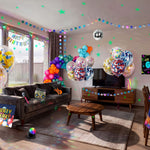Complete kids glow disco birthday party decorations filling a living room, including confetti balloons, multicolour balloon garland, dot garland, glow star wall decorations, disco ball, disco ball, neon backdrop, table cover, microphone and boombox props from a OneStopBox all-in-one party kit.