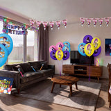 Living room decorated for a K-Pop Demon Hunters themed kids birthday party with character printed balloons in pink, blue, yellow and purple, triangle bunting, happy birthday banner, backdrop, microphone props and themed table cover from a OneStopBox party decorations kit.