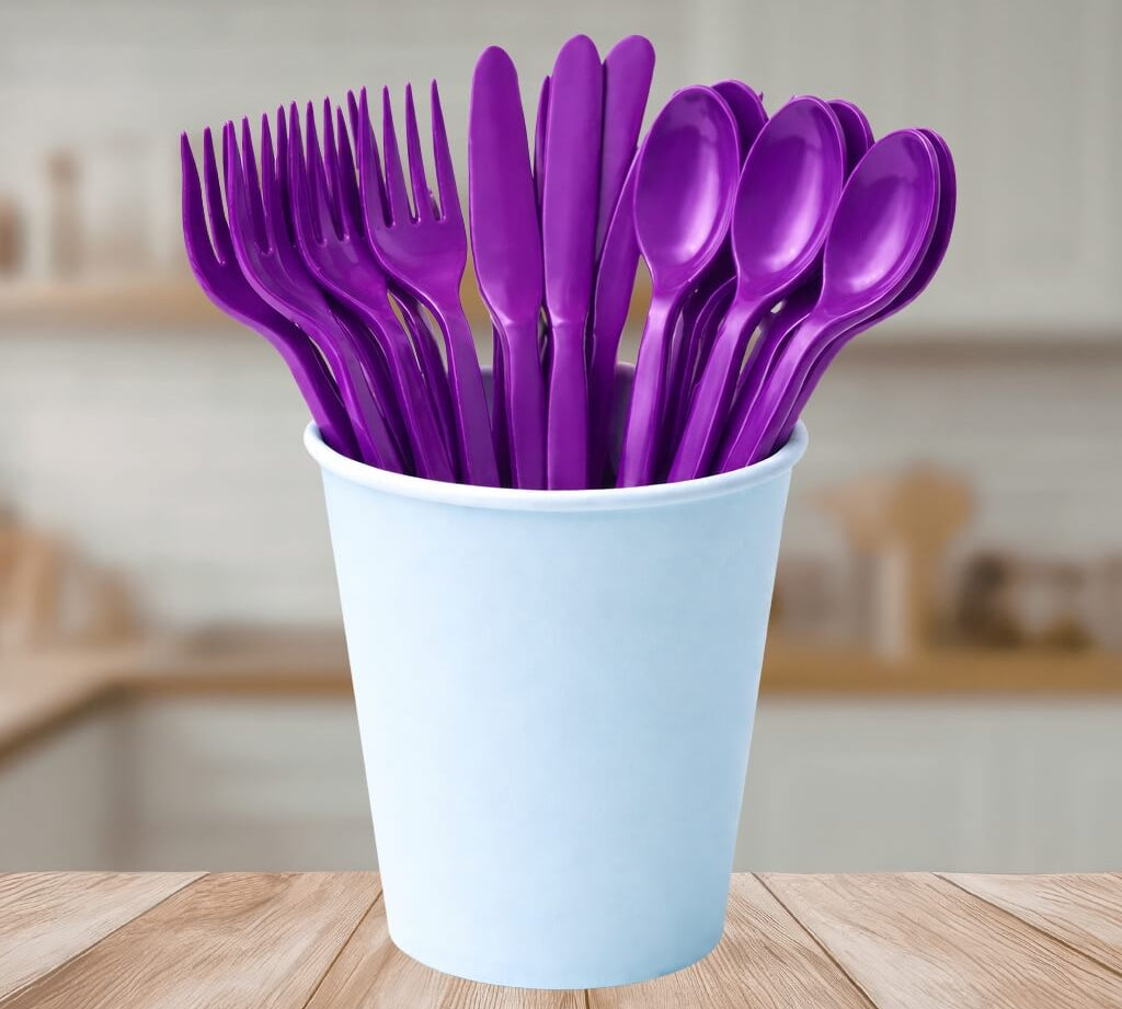 Purple reusable cutlery set including forks, knives, and spoons in a white cup on a wooden table.