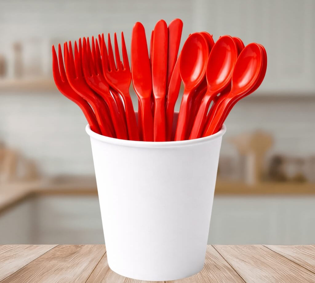 Red reusable plastic cutlery including forks, knives, and spoons in a white cup on a wooden table.
