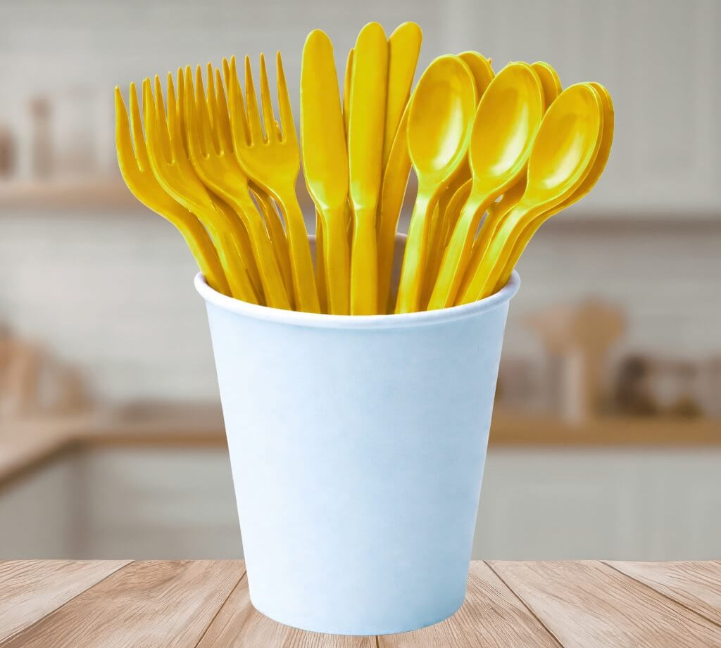 Yellow reusable plastic cutlery set in a white cup on a wooden table with a blurred kitchen background.